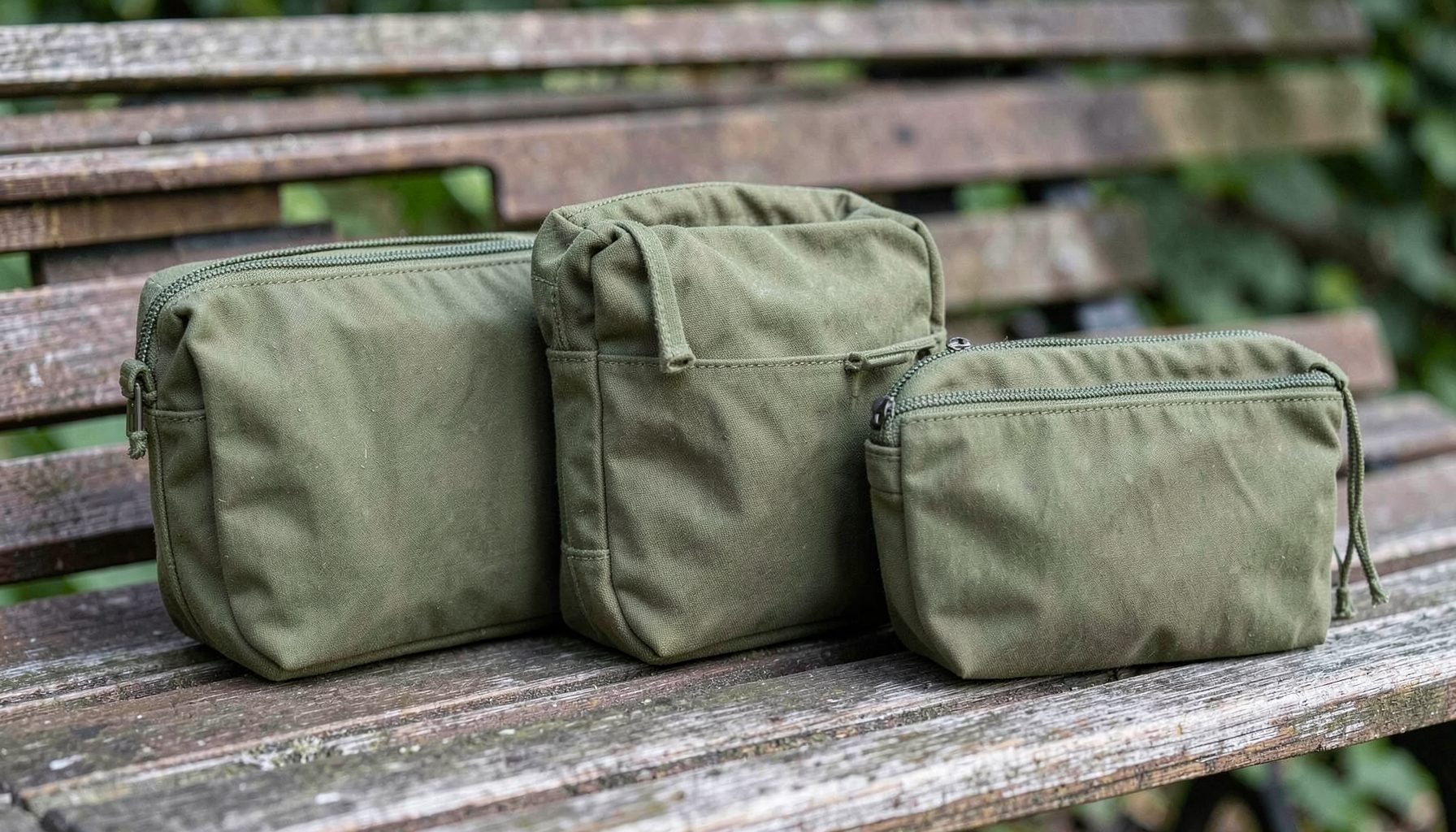 A coordinated matching set of three waxed canvas bags in matching olive tones — a dopp kit, razor pouch, and makeup pouch — grouped together on a wooden bench.