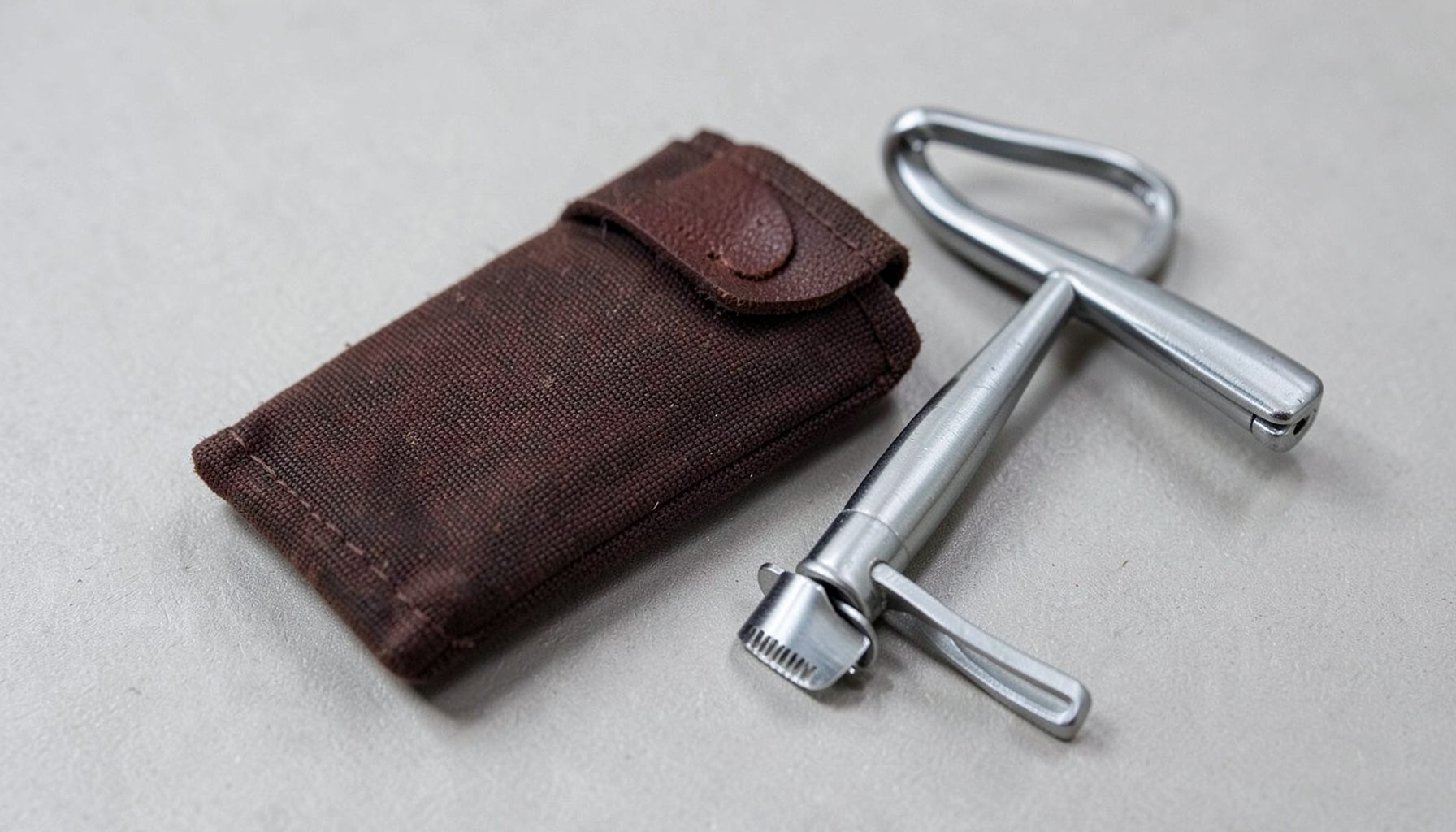 A slim razor pouch in dark brown waxed canvas with a single leather pull tab, lying flat beside a safety razor.