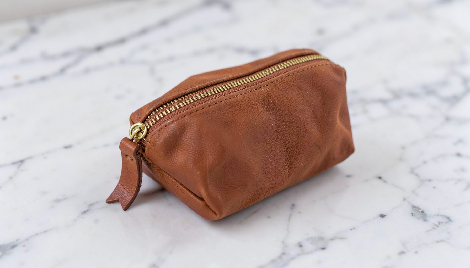 A small makeup pouch in warm brown waxed canvas with a brass zipper and leather pull, sitting on a marble surface.