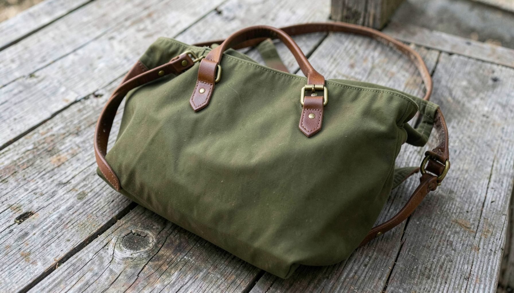 A waxed canvas tote handbag with rich brown leather straps and brass hardware, resting on a weathered wooden surface in soft natural light. The canvas has a deep olive-green tone with subtle waxed texture.