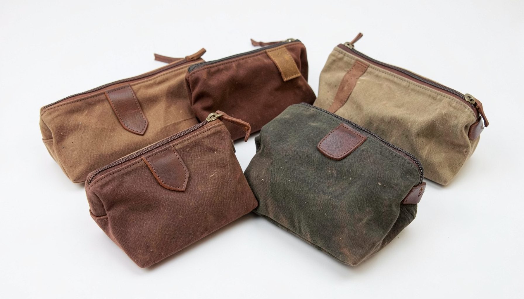 Several waxed canvas makeup pouches in earthy tones with leather accents, arranged on a clean white surface